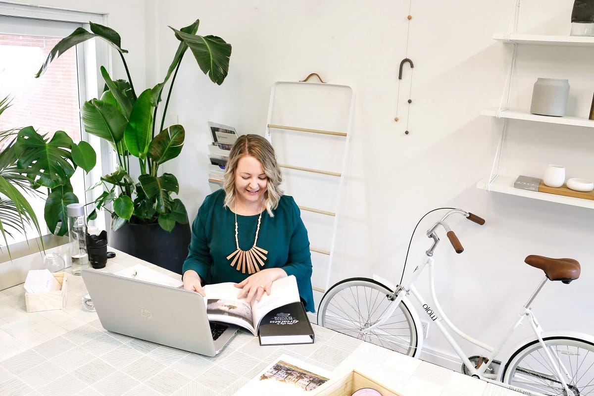 Business_consulting_services_home_header.1 Women in a green top sitting at a desk reading a book. On the desk is a laptop. There is a bike and green plants in the background.