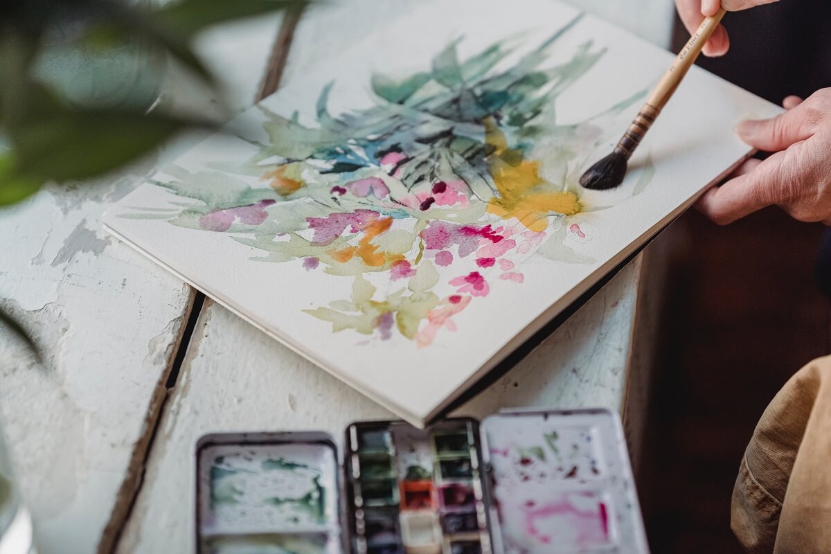 cash-flow-header There is a watercolour painting of flowers in bright colours with a paintbrush being held by a person. In the foreground is half a watercolour palette.